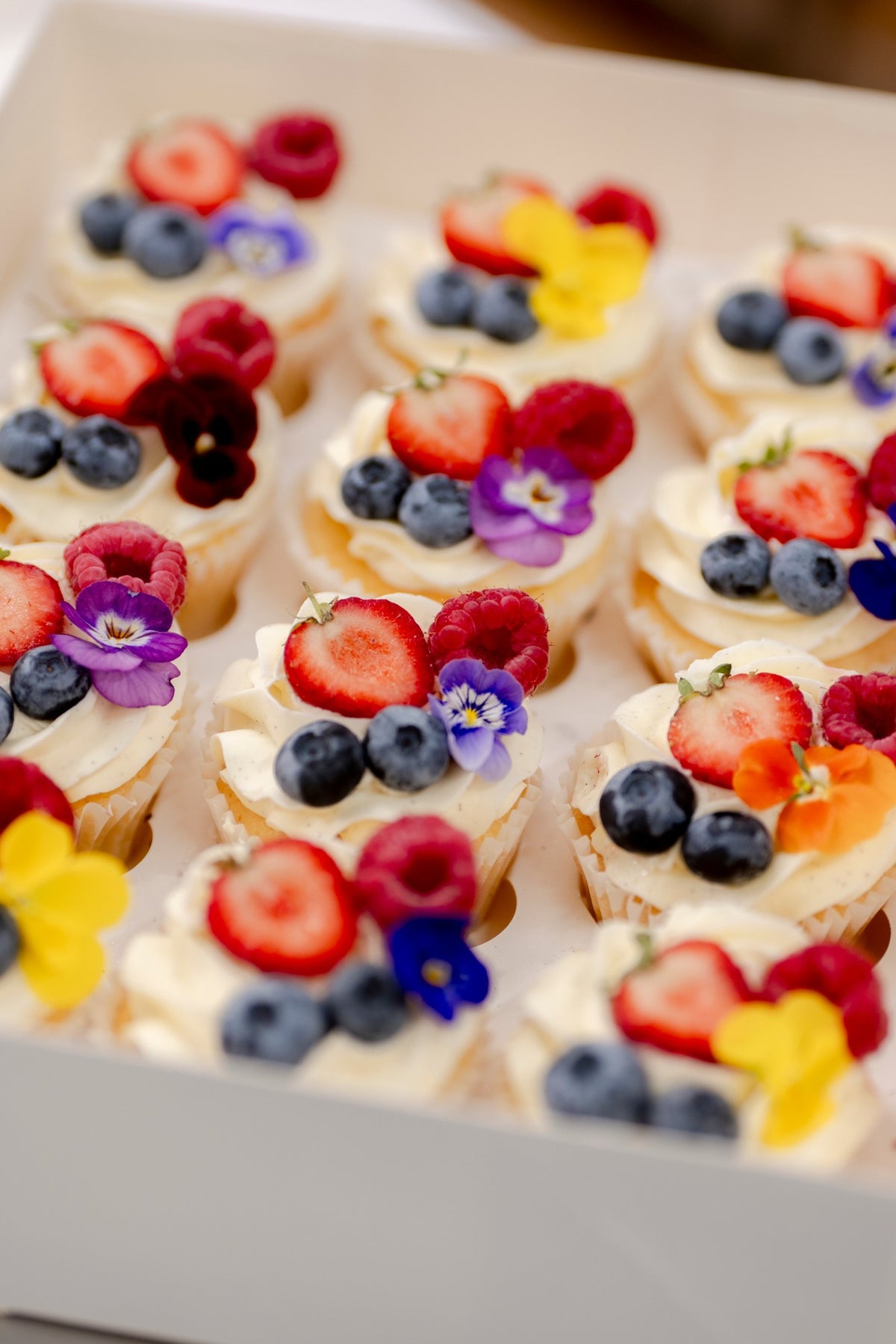 A box of vanilla cupcakes topped with fresh berries and edible flowers